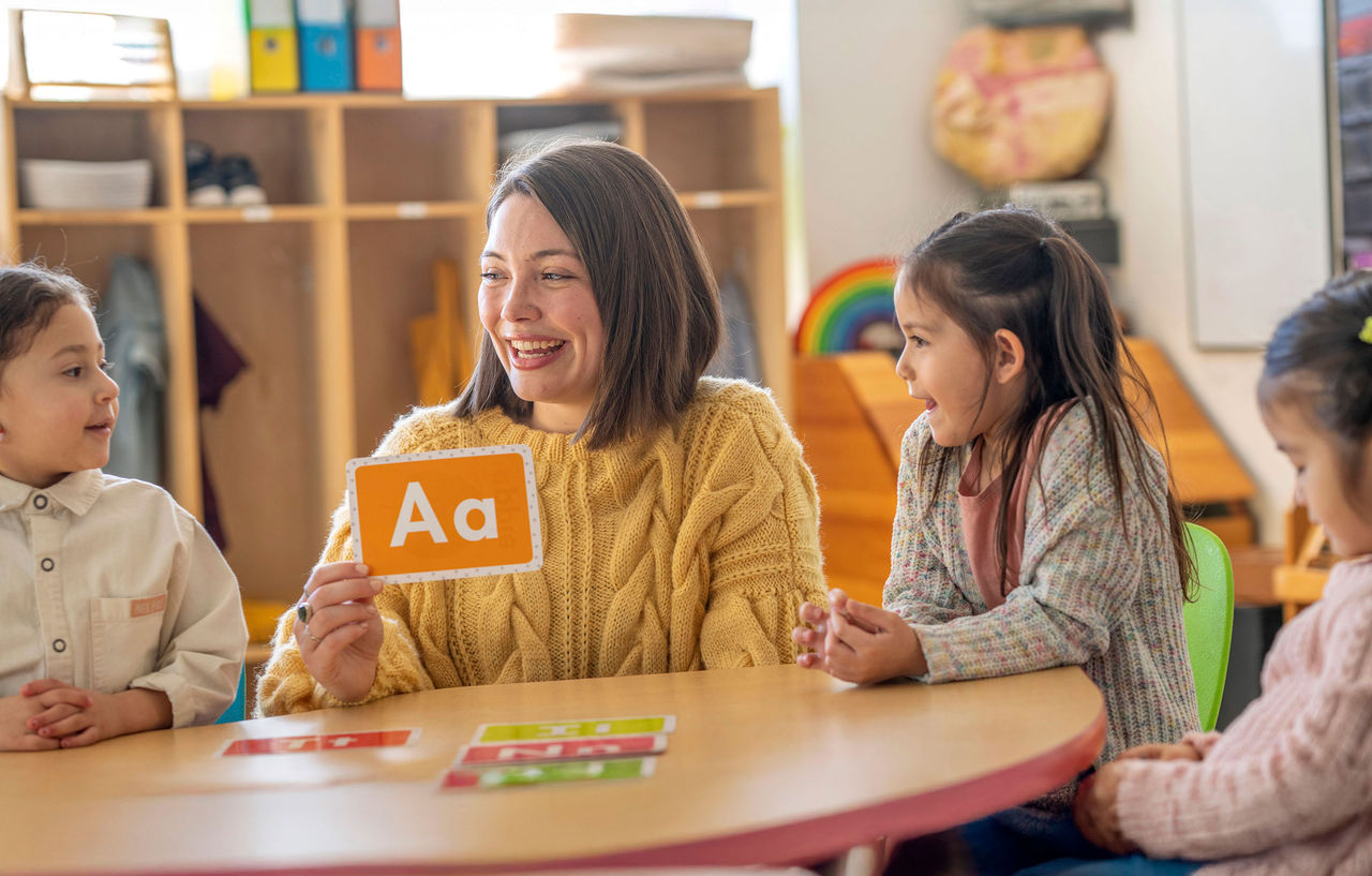 A Kindergarten teacher sits with a small group of students as she teaches them the different sounds each letter of the alphabet makes. Each of the students are dressed casually and paying close attention to their teacher.