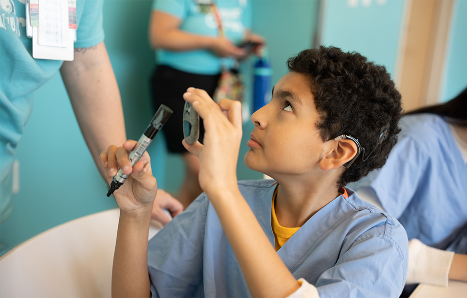 Child wearing scrubs holding a cochlear implant in one hand and a marker in the other