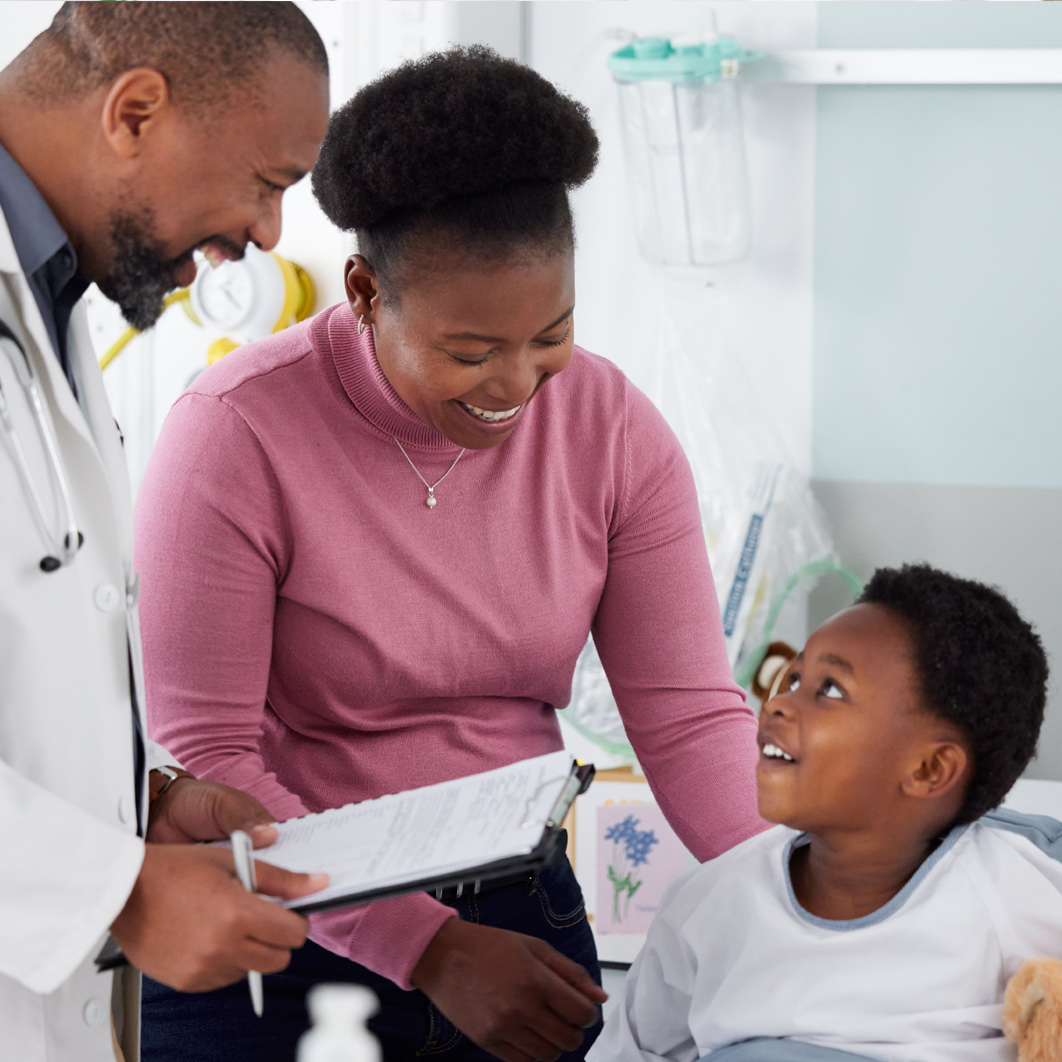 African American doctor holding a clipboard at the beside of a child and his mother.