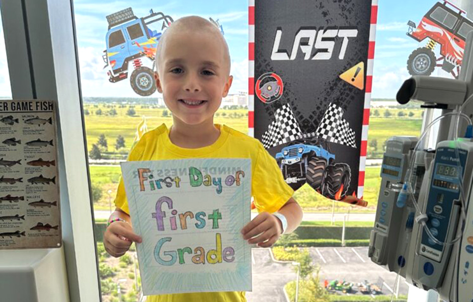 Porter, a lymphoma patient, holds a first day of first grade sign in front of a window with monster truck decals.