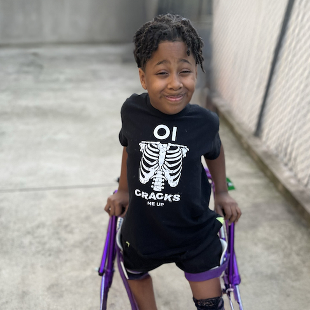 Child wearing OI Cracks Me Up shirt, standing and smiling with their purple walking brace