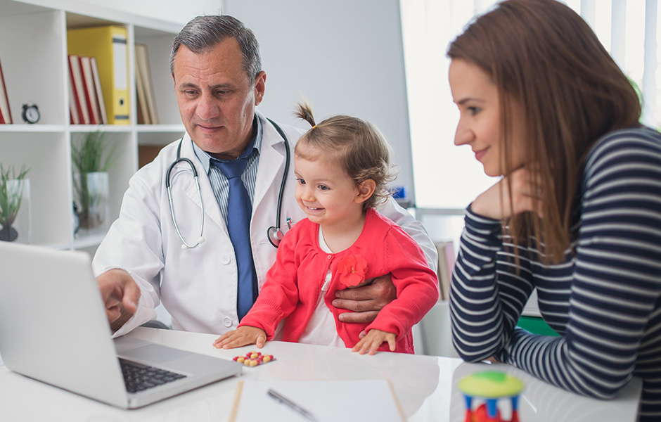 Male doctor holds toddler girl as they look at laptop with mother
