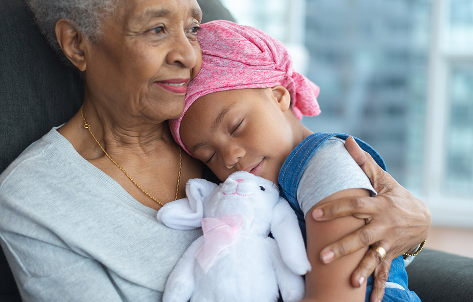 Grandmother holds sleeping child in a chair, child wears head covering and has a stuffed animal